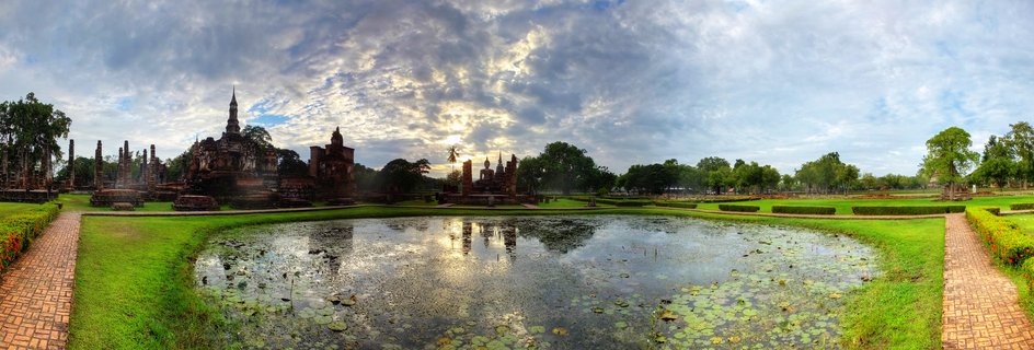 Ancient Buddhist temple ruins with tall chedi stupas reflected in a lotus-covered pond surrounded by green lawns and brick pathways under dramatic cloudy sky at Sukhothai Historical Park, Thailand
