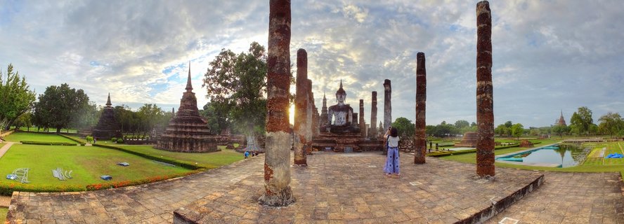 Panoramic view of ancient Buddhist temple ruins with large seated Buddha statue centered among tall weathered brick columns, green lawns, and visitor at Sukhothai Historical Park in Thailand