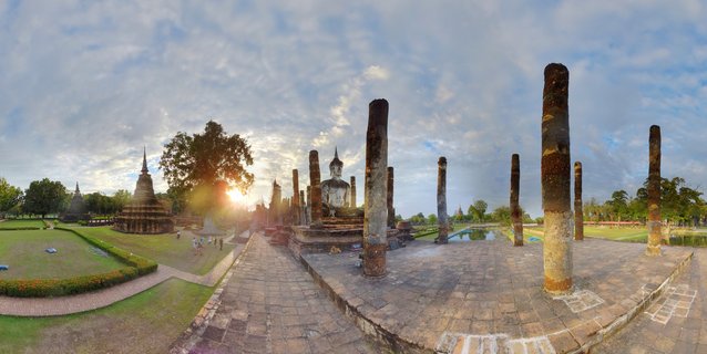 Panoramic view of ancient Buddhist temple ruins with a large seated Buddha statue, weathered stone pillars, and brick stupas at Sukhothai Historical Park in Thailand during sunset with visitors walking on pathways