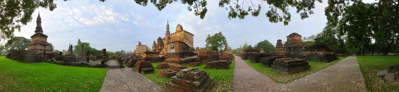 Panoramic view of ancient brick Buddhist temple ruins with multiple stupas and chedis at Sukhothai Historical Park in Thailand, with green lawns, brick pathways, and trees under a blue sky with scattered clouds.