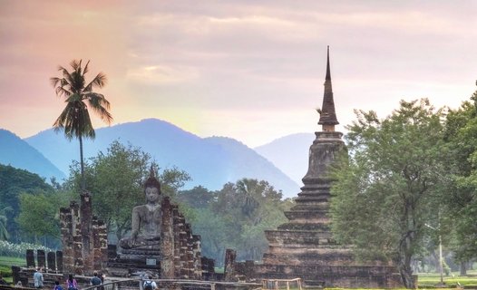 Ancient seated Buddha statue and brick stupa temple ruins at Sukhothai Historical Park in Thailand with palm trees, mountains in background, and pink-orange sunset sky