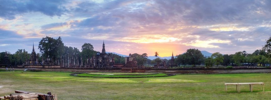 Panoramic view of ancient Buddhist temple ruins with multiple pointed stupas and chedis at Sukhothai Historical Park in Thailand during golden hour sunset with dramatic cloudy sky and green lawn foreground