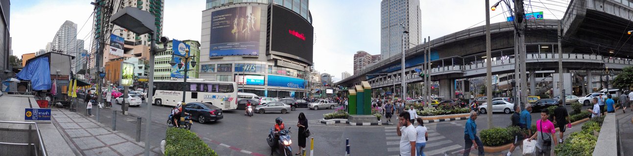 Wide street view of busy Bangkok intersection with elevated train tracks, multiple vehicles including motorcycles and buses, pedestrians crossing streets, and tall commercial buildings with billboards