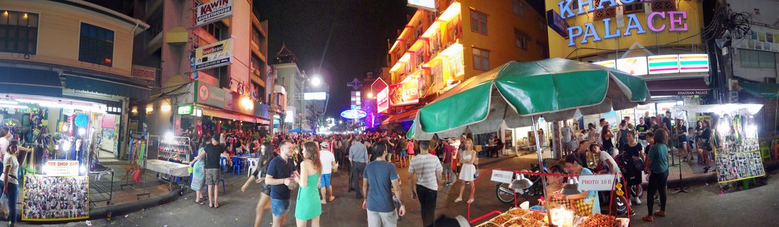 Busy night street scene showing crowds of tourists walking past illuminated vendor stalls and neon-lit buildings on Khao San Road in Bangkok
