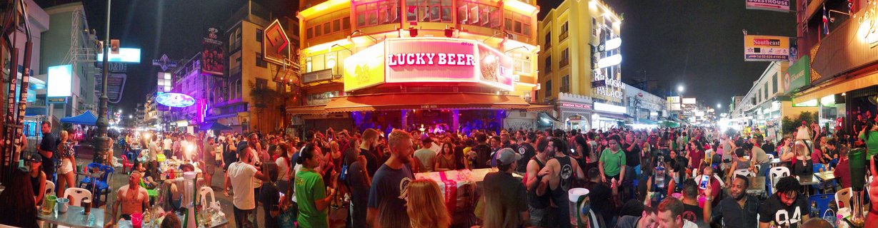 Crowded street scene at night showing hundreds of people walking and dining at outdoor tables on Khao San Road in Bangkok, with bright neon signs including Lucky Beer on yellow and white buildings