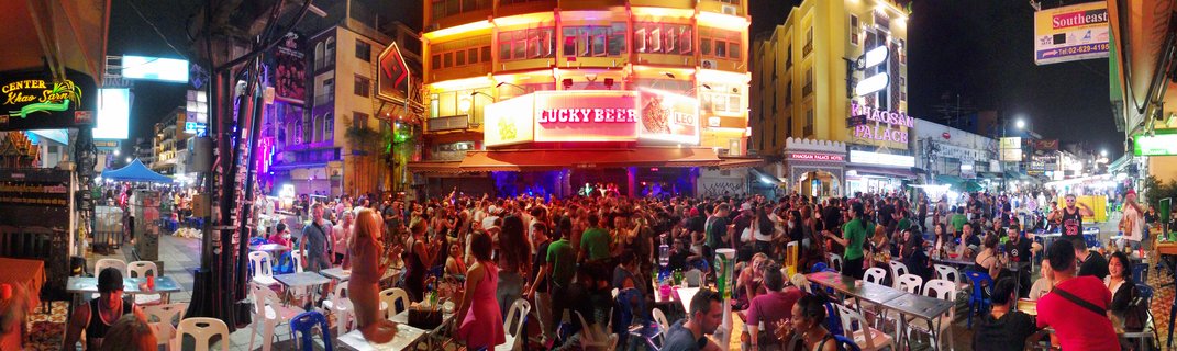 Crowded street scene at night on Khaosan Road Bangkok with neon signs, outdoor bars, tourists dining at plastic tables, and multi-story buildings with illuminated signage