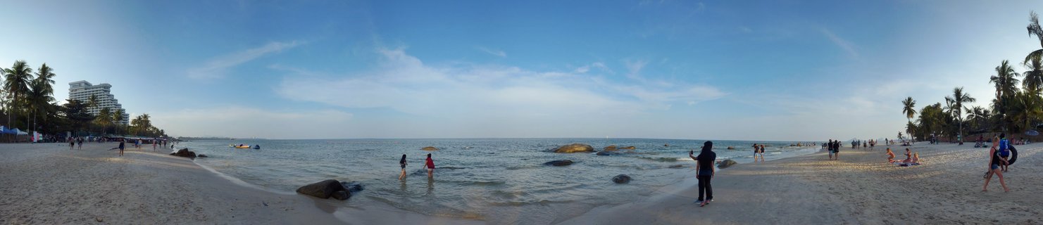 Wide panoramic beach scene showing people on sand and in shallow ocean water, with palm trees along the shoreline, rocks in the water, and a hotel building visible on the left side under a blue sky