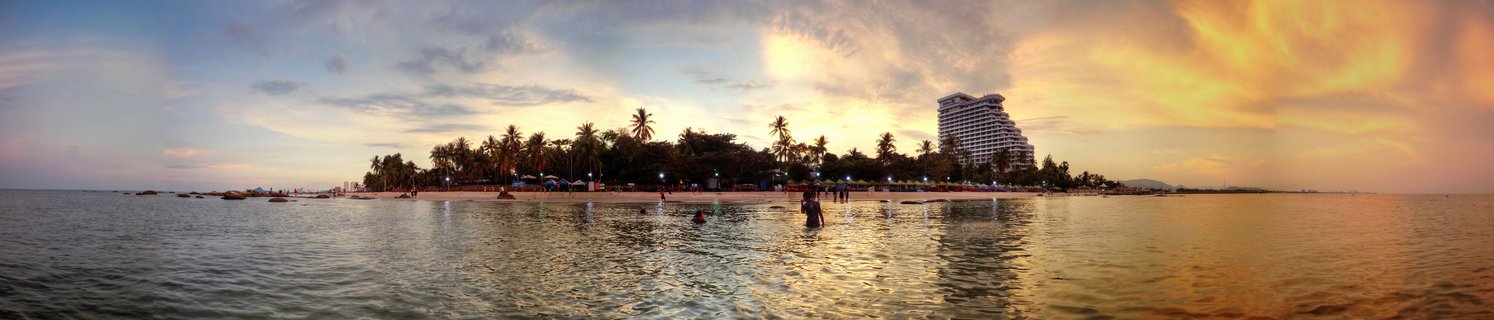 Panoramic view of Hua Hin Beach at sunset showing people swimming in calm water, palm trees along the shore, and a large white hotel building against a golden sky