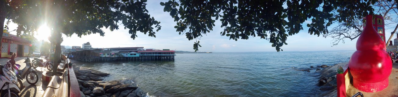 Coastal view showing stilt houses over water, red decorative sculpture on right, motorcycles parked on left, tree branches overhead, and calm ocean extending to horizon