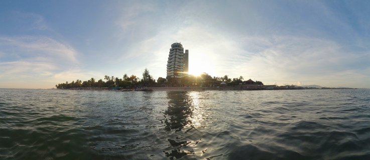 A tall hotel building on a beach at sunset with palm trees, viewed from the water with sun reflection on the sea surface