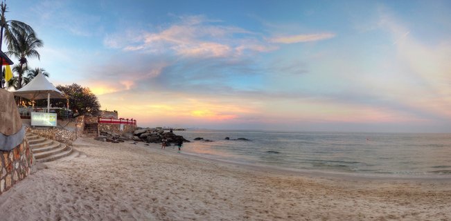 Panoramic beach scene at sunset showing sandy shore, calm ocean waters, colorful sky with pink and orange clouds, palm trees, white tent structure with stairs, rocks along shoreline, and small figures of people walking near the water