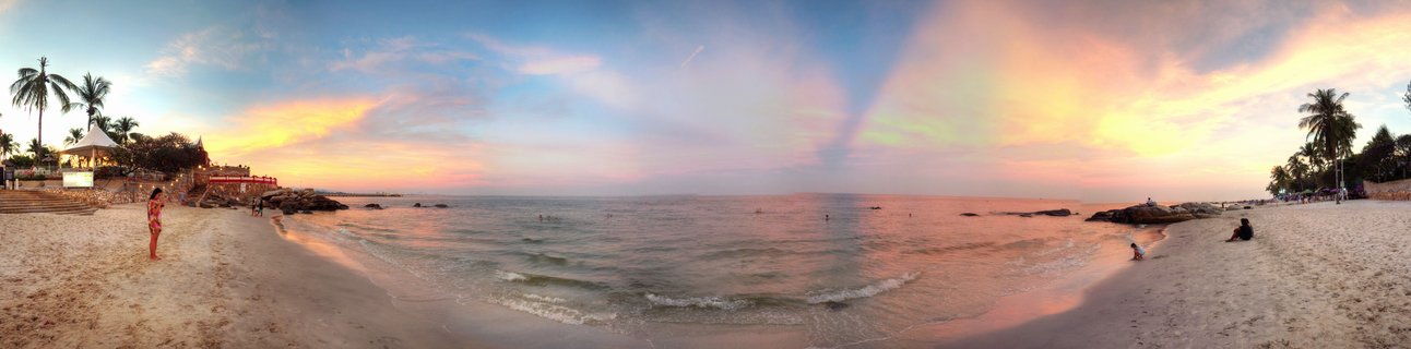 Panoramic beach scene at sunset showing sand, ocean waves, palm trees on both sides, colorful sky with orange and pink clouds, and people on the shoreline