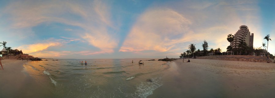 Panoramic beach scene at sunset in Hua Hin, Thailand showing sand, ocean waves, people swimming, palm trees, and a large resort hotel building under colorful cloudy sky