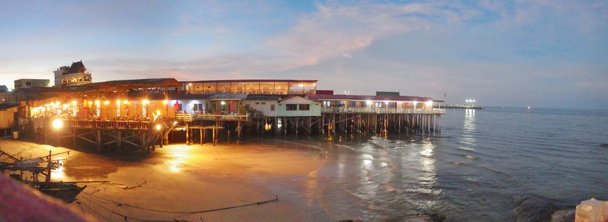 Panoramic view of illuminated restaurant buildings on wooden stilts over water at Hua Hin beach during twilight, with warm lights reflecting on wet sand and calm sea