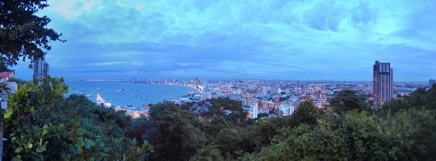 Aerial view of Pattaya city coastline at dusk with illuminated buildings, boats in the bay, and tropical trees in foreground under cloudy blue sky