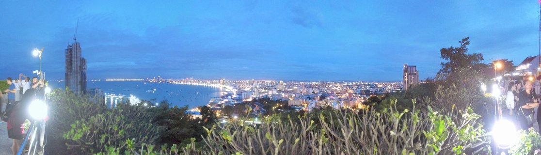 Panoramic view of Pattaya city and bay at dusk with illuminated buildings along the coastline, tourists at viewpoint in foreground, deep blue twilight sky