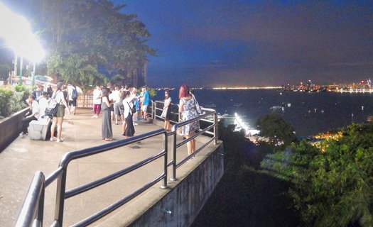 Group of tourists on elevated viewing platform at night overlooking illuminated Pattaya Bay with city lights visible across the water in Thailand