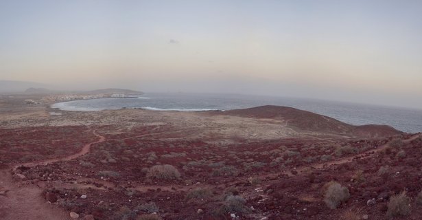 Reddish volcanic landscape with sparse vegetation overlooking a coastal bay town and ocean horizon in Tenerife, Spain
