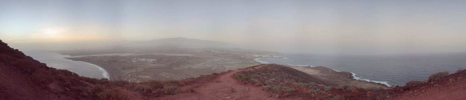 Panoramic landscape showing reddish volcanic terrain overlooking El Médano coastal bay at sunset with Mount Teide visible in atmospheric haze, Tenerife, Canary Islands, Spain