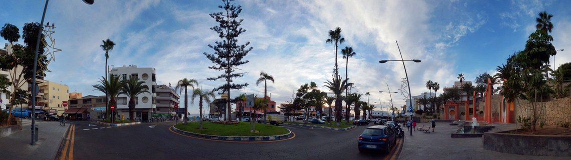 Panoramic street view of a roundabout with palm trees, landscaped green space, white buildings, and pedestrians in Los Cristianos, Tenerife under a blue sky with scattered clouds