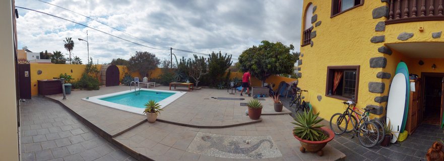 A rectangular swimming pool with turquoise water in a stone-paved courtyard surrounded by a yellow stucco building with volcanic stone trim. Two bicycles and a blue-and-white surfboard lean against the building. Potted succulents and agave plants are scattered around the patio, which features an embedded mosaic lizard design. A person in a red shirt walks in the background near a wooden gate. Palm trees and overcast sky visible beyond the yellow boundary wall.