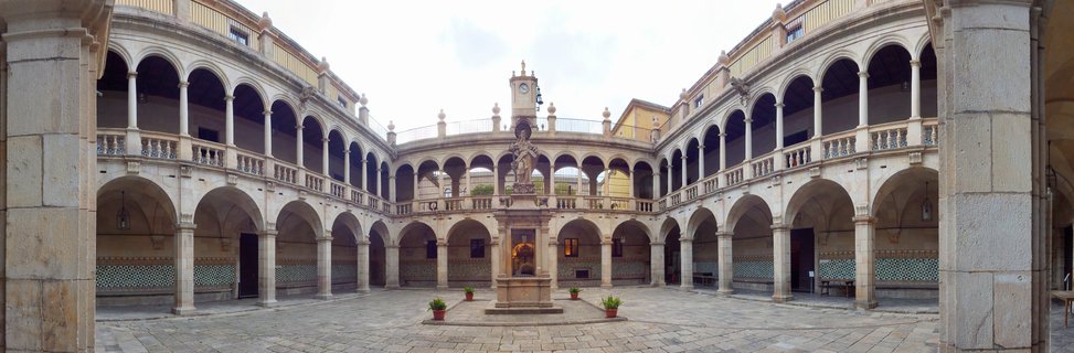 Historic stone courtyard with two levels of arched colonnades surrounding a central fountain with statue, potted plants, and decorative tilework on walls