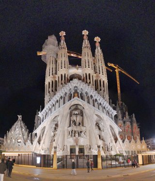 Night photograph of Sagrada Familia basilica in Barcelona with illuminated white stone facade, tall spires with decorative crowns, construction cranes visible, people walking on street in foreground against dark sky