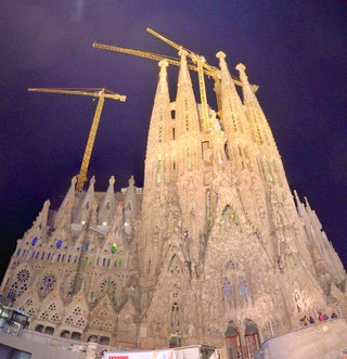 Night photograph of Sagrada Familia basilica in Barcelona showing illuminated stone spires, colorful stained glass windows, and yellow construction cranes against a dark blue sky