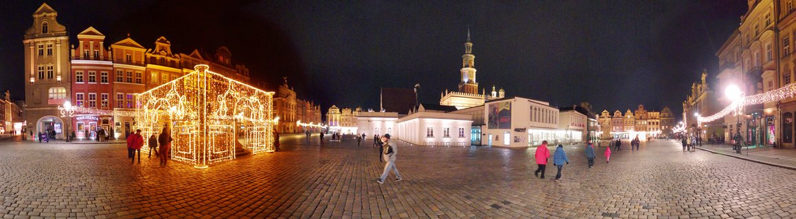 Panoramic night view of Poznań's Old Market Square showing a large golden Christmas light installation, surrounded by illuminated Renaissance and Baroque buildings including the Town Hall tower, with pedestrians walking on cobblestone pavement