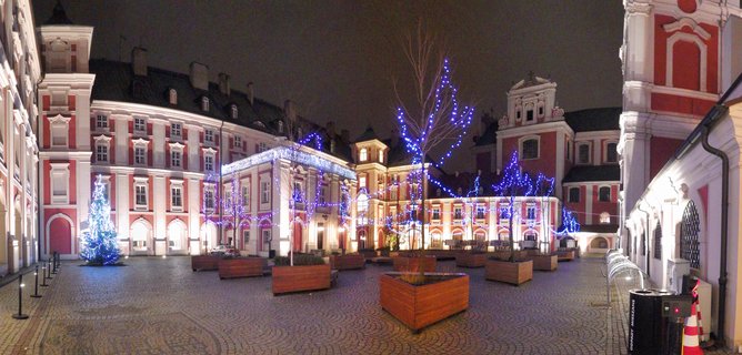 Night view of a historic courtyard with red and white baroque buildings decorated with blue Christmas lights, cobblestone ground, illuminated trees in wooden planters