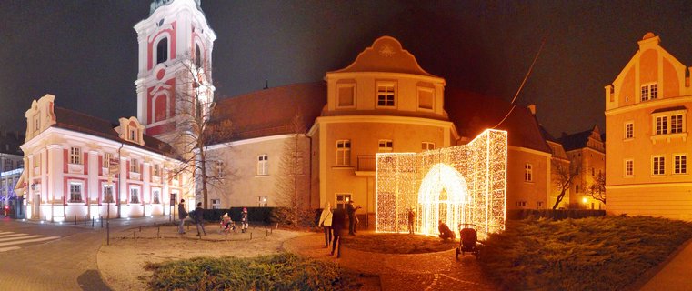 Night panorama of historic Poznan square with illuminated baroque church tower, warm yellow buildings, and glowing light sculpture archway with people walking