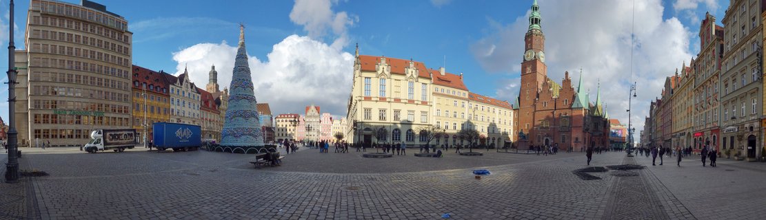 Wide panoramic view of Wrocław Market Square showing historic colorful buildings, Gothic Town Hall tower with green spire, large blue Christmas tree structure, and people walking on cobblestone pavement under blue sky with clouds