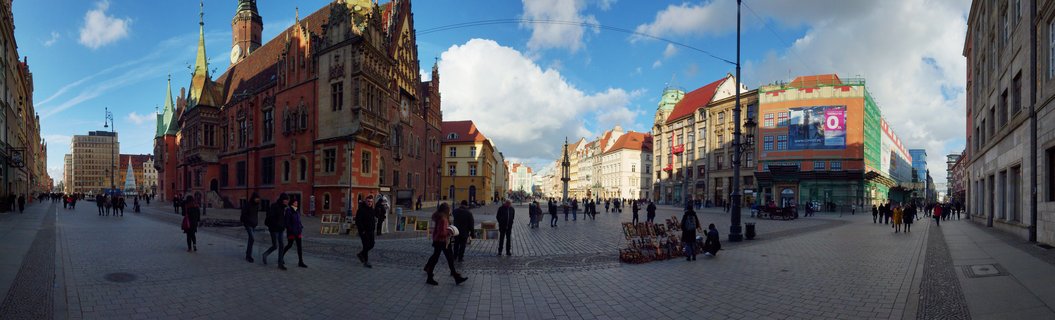 Wide view of Wrocław's Market Square showing the Gothic Town Hall with twin towers, surrounded by colorful historic tenement buildings, pedestrians on cobblestone pavement, street vendor art display, blue sky with clouds in winter.