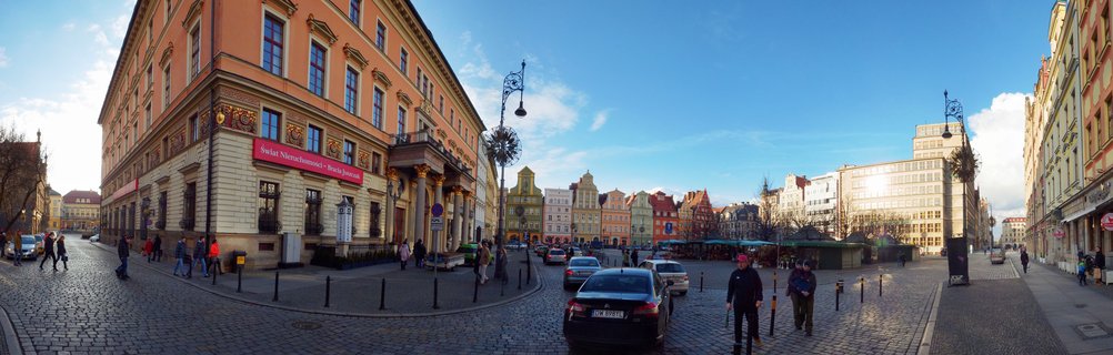 Panoramic street view of Wroclaw's Old Town showing colorful historic buildings, cobblestone streets, pedestrians, and cars under a blue winter sky