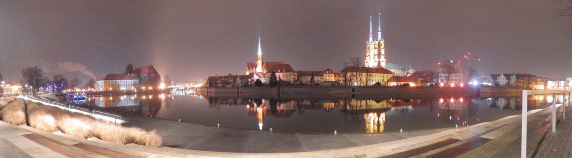 Night panoramic view of Wroclaw's Cathedral Island showing illuminated Gothic church spires and historic buildings reflected in the Odra River, with a modern riverside walkway in the foreground