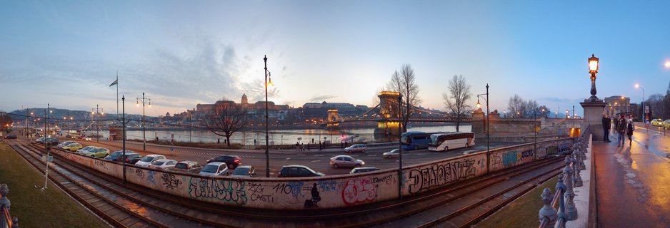 Panoramic view of Budapest's Chain Bridge spanning the Danube River at dusk with Buda Castle illuminated on the hill, tram tracks in foreground, wet pavement reflecting street lights