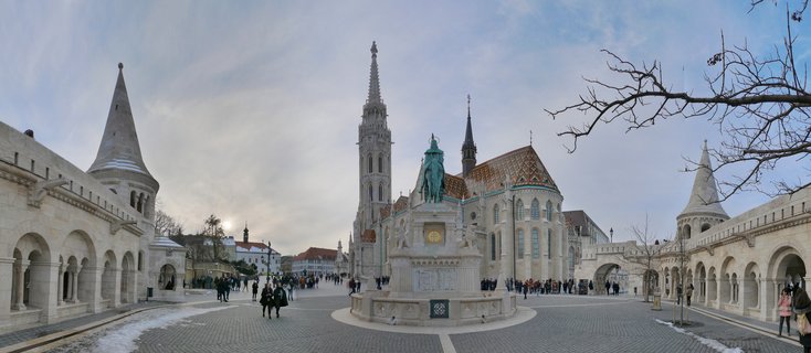 Wide view of Fisherman's Bastion's white stone towers with conical roofs flanking Matthias Church's colorful tiled roof and tall Gothic spire in Budapest, with tourists walking on cobblestone plaza and statue monument in foreground