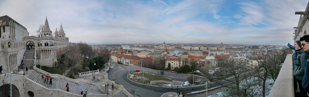 Panoramic view from Fisherman's Bastion showing white stone towers, stone stairways with tourists, Budapest cityscape with red-roofed buildings, Danube River, and Hungarian Parliament dome in the distance under partly cloudy winter sky