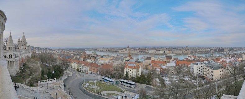 Panoramic cityscape of Budapest showing Fisherman's Bastion towers on the left, Danube River through the center, Hungarian Parliament Building in the distance, and red-roofed buildings under a cloudy sky