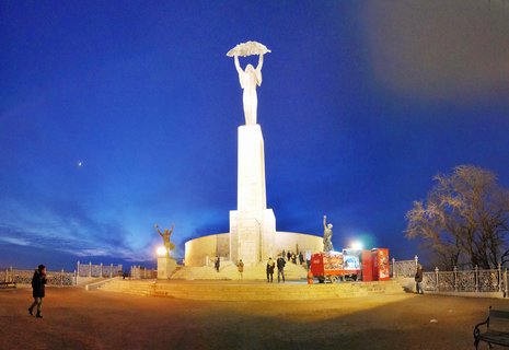 White limestone Liberty Statue holding palm leaf overhead, illuminated at twilight on Gellért Hill in Budapest with tourists at base and deep blue sky with crescent moon