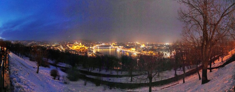 Panoramic night photograph of Budapest showing illuminated Buda Castle, Chain Bridge, and Parliament Building along the Danube River with snow-covered foreground and bare trees at blue hour
