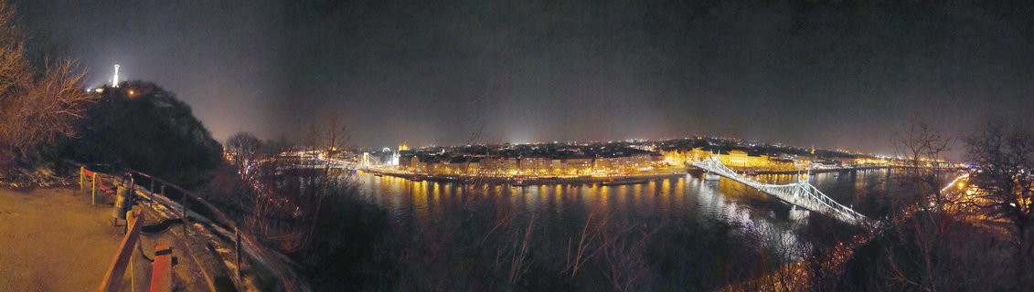 Night panoramic view of Budapest showing the illuminated Chain Bridge crossing the Danube River with city lights reflecting on water and the Liberty Statue visible on Gellért Hill