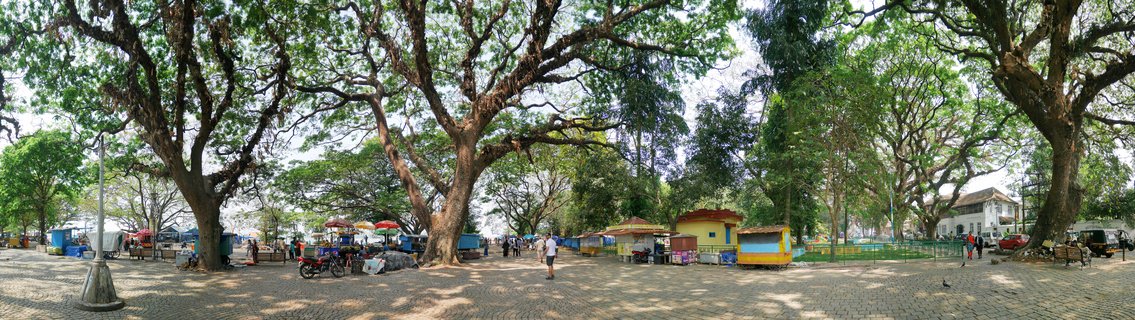 Wide panoramic view of a public square in Fort Kochi with large banyan trees, colorful vendor stalls, cobblestone ground, people walking, parked motorcycles, and colonial buildings