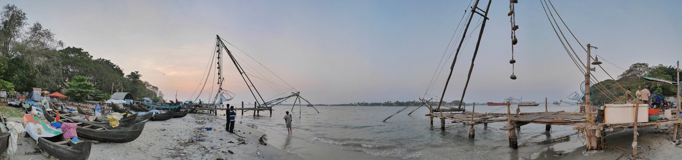 Panoramic view of multiple large Chinese fishing nets along a beach shoreline at dusk in Kochi, India, with fishing boats on the sand and people standing near the water
