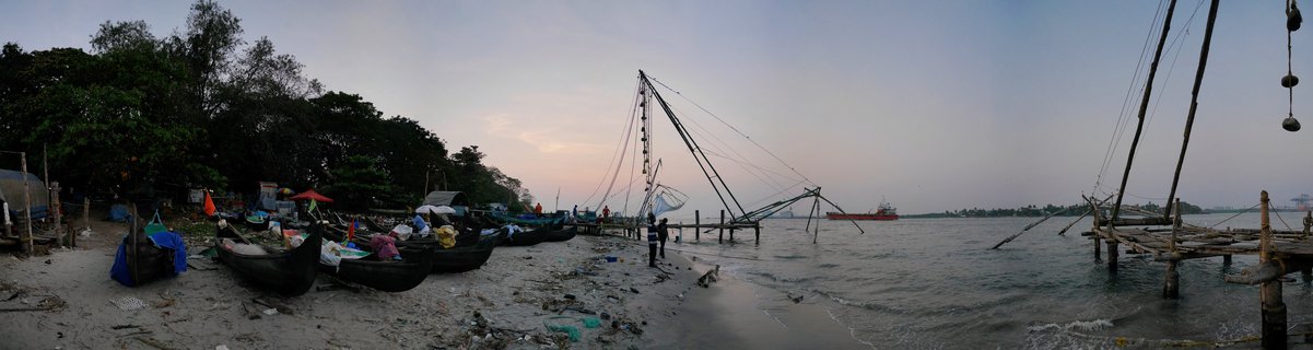 Traditional Chinese fishing nets on wooden frames along Kochi harbor shoreline with beached fishing boats, trees, and distant cargo ship under twilight sky