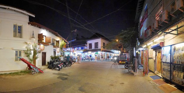 Night panoramic street view of Fort Kochi showing white colonial buildings on the left, illuminated jewelry and textile shops on the right, pedestrians walking, parked motorcycles, an auto-rickshaw, and overhead electrical wires against a dark sky