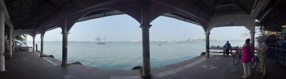 Panoramic view from under a covered pier with arched concrete columns overlooking the Arabian Sea, with fishing boats visible on the water and people standing on the right side of the jetty structure