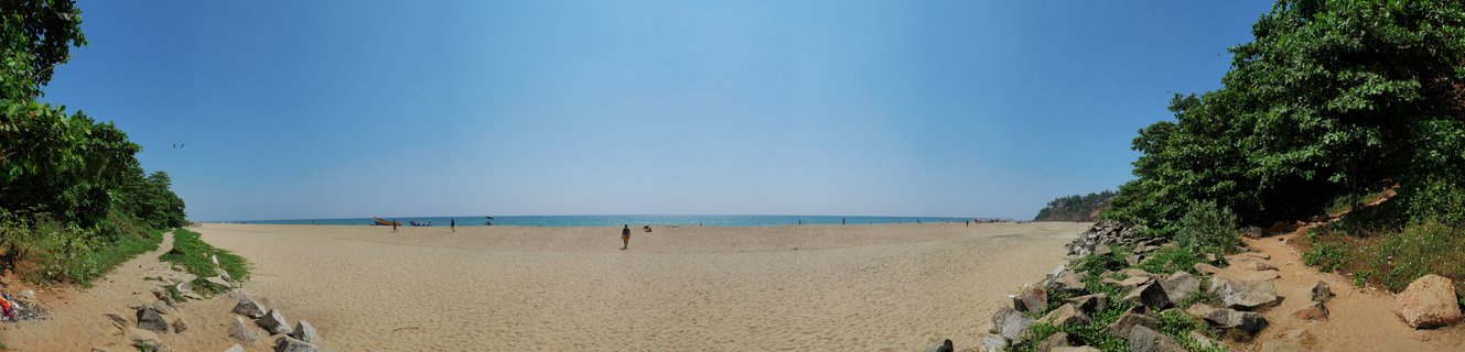 Wide panoramic photograph of Varkala Beach in Kerala, India showing expansive sandy beach, calm blue Arabian Sea on the horizon, clear sky, and lush green trees and rocks on both sides with a few people walking on the sand