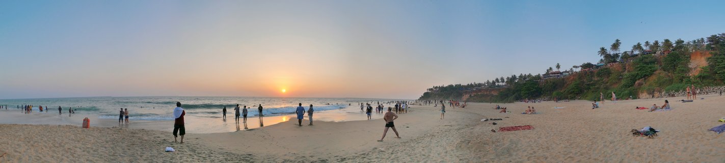 Wide panoramic view of Varkala Beach at sunset showing people on sand, ocean waves, setting sun on horizon, and red cliffs with palm trees on the right side