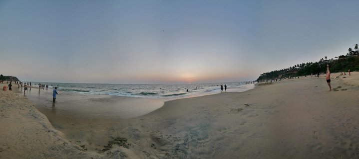 Wide beach scene at sunset showing people walking on wet sand, ocean waves, palm trees on a cliff hillside, and the sun setting on the horizon over the Arabian Sea in Varkala, India